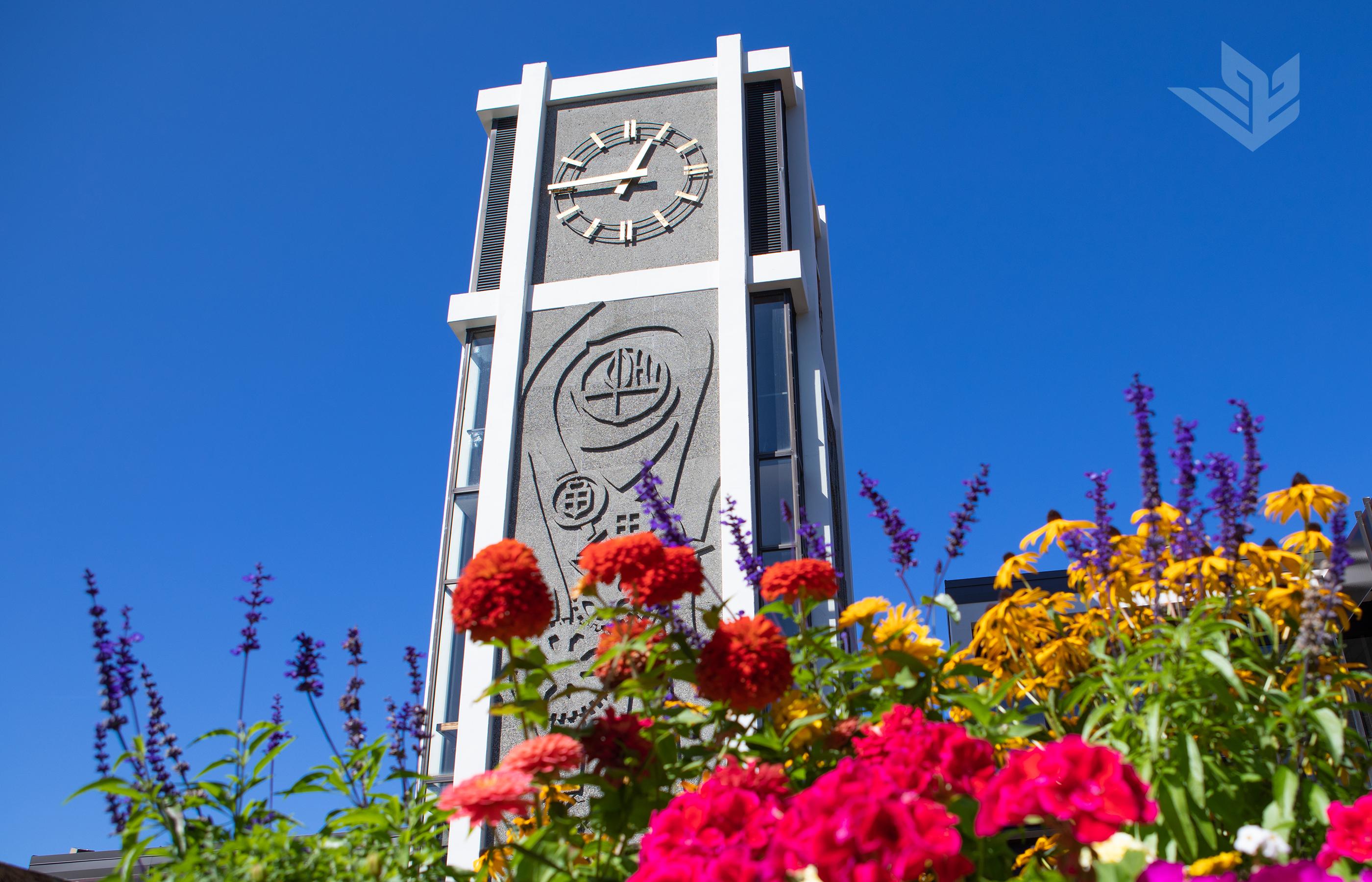 The iconic Demaray Hall clocktower behind summer flowers and in front of a cloudless blue sky