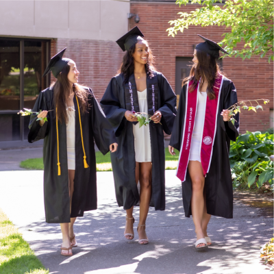 Three  smiling female SPU graduates walking on campus while wearing their caps and gowns
